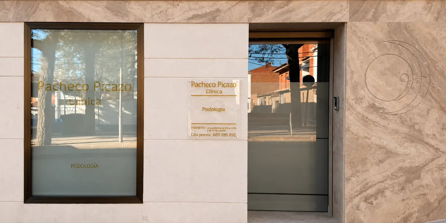 Fachada de la clínica en piedra clara y travertino: escaparate con el nombre del centro, placa con la especialidad de podología y el horario, puerta de entrada acristalada y el símbolo de la clínica grabado en la piedra